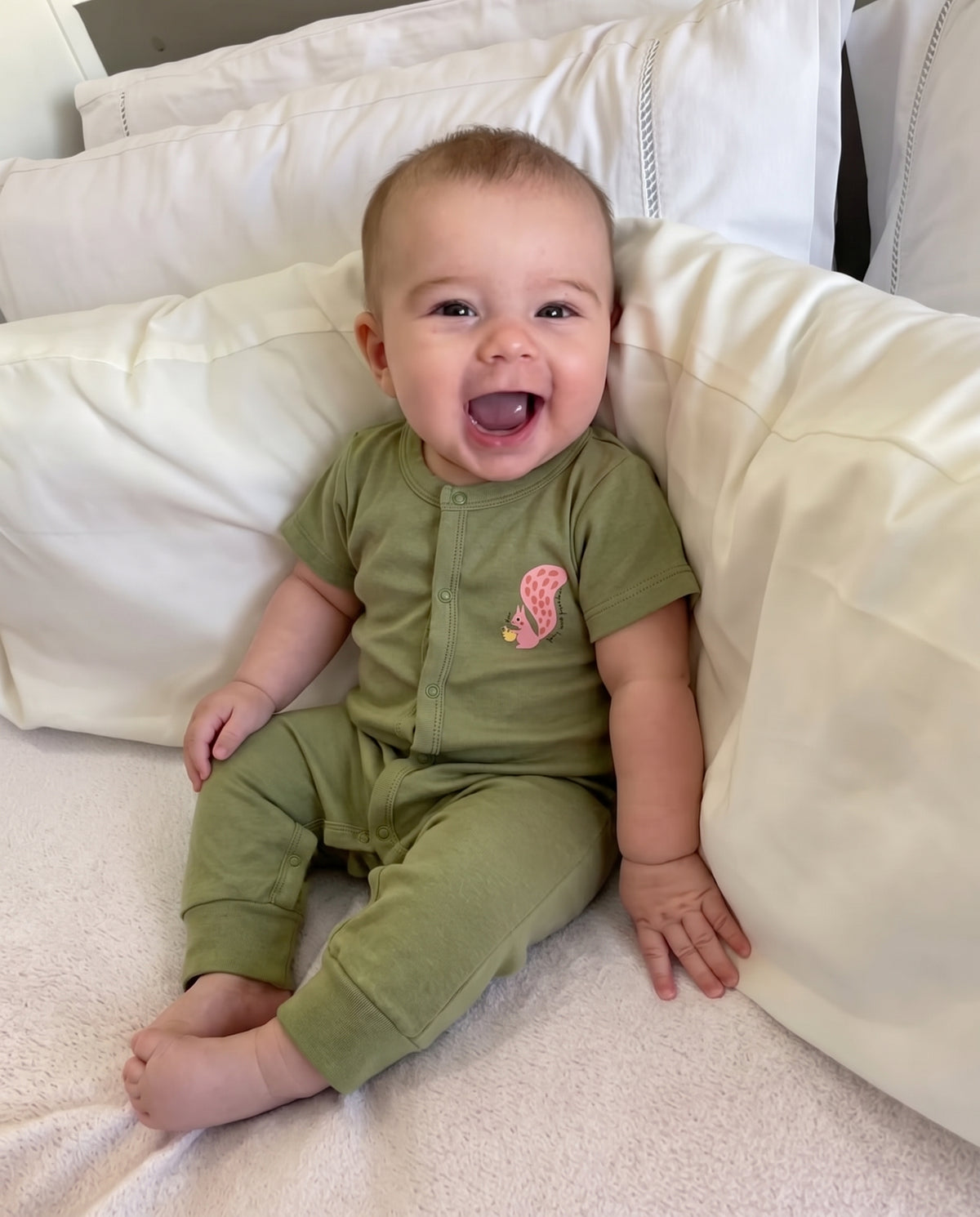 Baby sitting on a bed wearing the olive-green Olive Grove short-sleeve jumpsuit